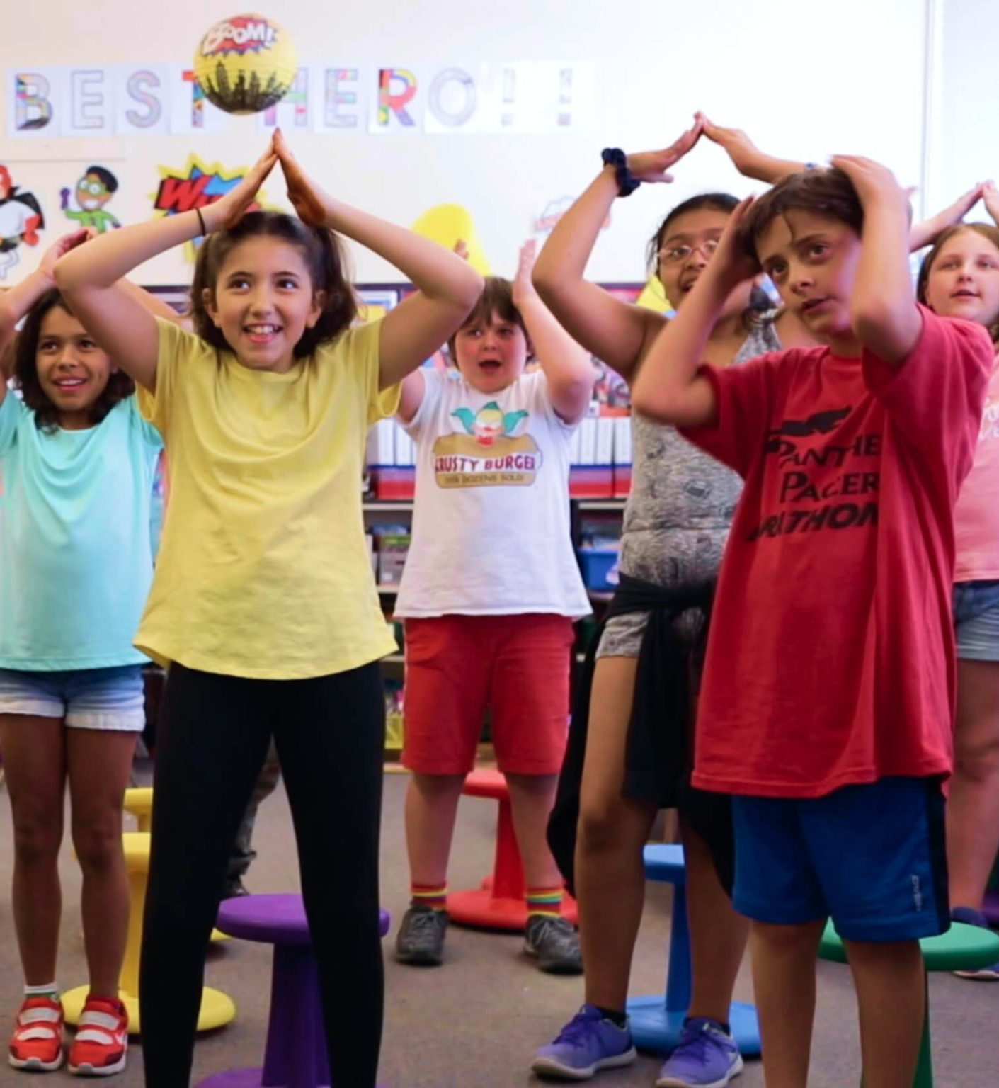 A group of kids in a classroom raise their arms over their heads and bring their hands together in a point.
