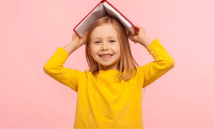 Young girl smiles at the camera while holding an open book over her head.